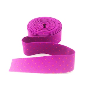 May include: A roll of vibrant magenta fabric ribbon featuring a repeating pattern of small, evenly spaced orange polka dots. The ribbon is coiled into a tight cylinder at the top, with a single length extending forward in a gentle, flowing curve.