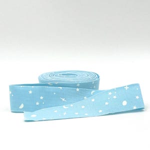 May include: A roll of light blue fabric ribbon featuring a repeating white pattern of small stars, crescent moons, and four-pointed sparkles. The ribbon is coiled into a flat circular shape with one end extending outward to display the print.