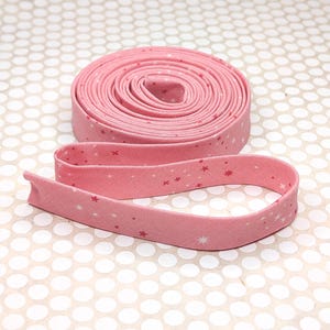 May include: A roll of pink fabric bias tape featuring a repeating pattern of small white and dark pink stars. The fabric tape is coiled into a circular shape with one end extending outward, displaying the consistent star print across the surface.