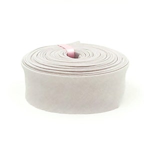 May include: A roll of light gray velvet ribbon wound into a circular shape. The fabric has a soft, matte texture and a consistent width throughout the spool. A small, thin pink paper band secures the end of the ribbon at the center of the roll.