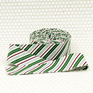 May include: A roll of fabric ribbon featuring a repeating diagonal striped pattern in shades of forest green, deep red, and thin white lines. The ribbon is wound into a tight circular spool with one end extending outward across the surface.