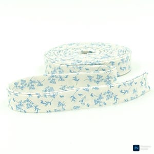 May include: A roll of off-white fabric ribbon featuring a repeating blue floral pattern. The ribbon is coiled into a flat circular shape with one end extending outward, displaying the continuous botanical print across the entire length of the trim.