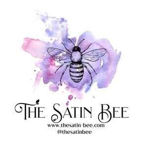 May include: A black ink illustration of a bee centered over a purple and pink watercolor splash. Below the bee, the text reads THE SATIN BEE in a decorative font, followed by the website www.thesatin-bee.com and the handle @thesatinbee.
