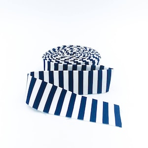 May include: A roll of fabric ribbon featuring a repeating vertical stripe pattern in dark navy blue and crisp white. The ribbon is coiled into a circular spool shape with one end extending outward, displaying the consistent striped design throughout.