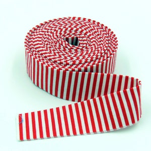 May include: A roll of fabric ribbon featuring a repeating pattern of vertical red and white stripes. The ribbon is coiled into a circular shape with one end extending outward, displaying the consistent striped design across the entire surface.