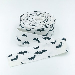 May include: A roll of white fabric ribbon with a repeating pattern of black bats. The ribbon is unrolled at the bottom, showing the bat design. The ribbon is likely for crafting or decorating.