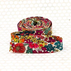 May include: A coiled length of fabric ribbon featuring a dense, multicolored floral pattern. The design includes vibrant red, orange, yellow, pink, and teal flowers and leaves set against a light cream background, creating a continuous botanical print.