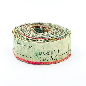 May include: A tightly coiled roll of vintage-style paper featuring a map design with muted green, beige, and red tones. The visible text on the outer edge includes the words DOMINI IC and MARCUS I. (U.-S.) printed in black ink along a dashed line.