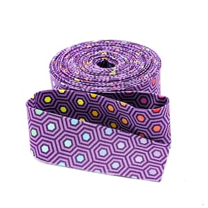 May include: A roll of purple fabric ribbon featuring a repeating geometric pattern of dark purple hexagons. The center of each hexagon contains a small, solid-colored dot in shades of yellow, orange, light blue, or pink, creating a vibrant contrast.