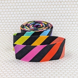 May include: A roll of fabric tape featuring a repeating diagonal stripe pattern in vibrant colors including black, yellow, orange, pink, and light blue. The tape is coiled into a circular shape with one end extending outward to display the pattern.