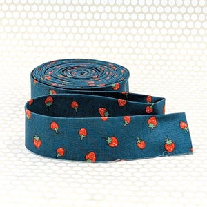 May include: A roll of blue fabric with a repeating pattern of red strawberries. The fabric is unrolled in the foreground, revealing the strawberry design. The background is a light beige color with a dotted pattern.