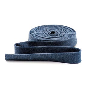 May include: A roll of blue denim fabric ribbon, with a length of the ribbon unrolled in the foreground. The fabric has a textured appearance and is a medium shade of blue. The ribbon is likely used for sewing or crafting.