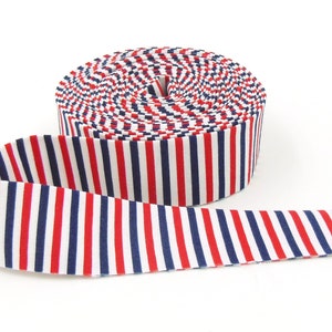 May include: A roll of fabric ribbon featuring a repeating vertical striped pattern in alternating colors of navy blue, white, and red. The ribbon is coiled into a circular spool shape with one end extending outward across the surface.