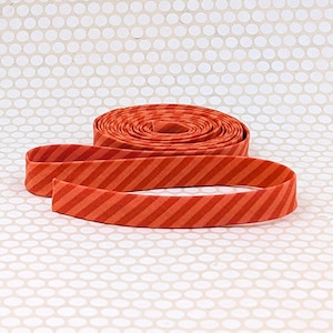 May include: A roll of fabric bias tape featuring a diagonal striped pattern in two shades of orange. The tape is coiled into a flat circular shape with a single length extending forward, displaying the consistent angled stripe design throughout.