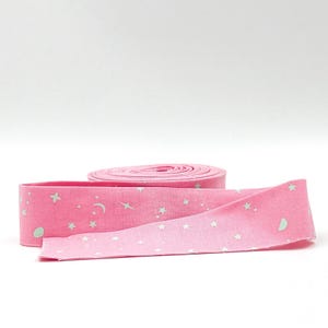 May include: A roll of pink fabric ribbon featuring a repeating pattern of small white stars, crescent moons, and four-pointed sparkles. The ribbon is coiled into a flat circular shape with one end extending outward to display the celestial print.
