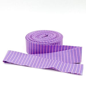May include: A roll of purple and pink striped fabric ribbon. The ribbon is rolled up and a length of the ribbon is laying out in front of the roll.