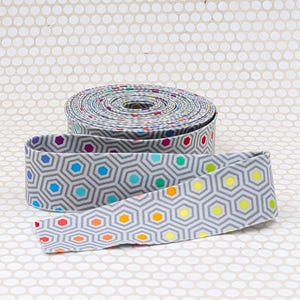 May include: A roll of gray fabric ribbon featuring a repeating geometric hexagon pattern. Each hexagon contains a small, solid-colored circle in shades of red, orange, yellow, green, blue, and purple, arranged in a continuous, flat-coiled spool.