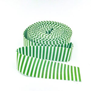 May include: A roll of fabric ribbon featuring a repeating pattern of vertical stripes in bright green and white. The ribbon is coiled into a circular shape with one end extending outward, displaying the consistent striped design throughout the material.