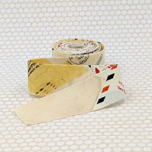 May include: A roll of fabric with a vintage postal theme. The fabric features a cream background with printed elements including stamps, text, and diamond patterns in red, black, and blue. The roll is partially unraveled, showing the fabric's design.