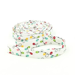 May include: A roll of white bias tape featuring a repeating pattern of colorful Christmas lights. The lights are red, green, yellow, and blue, connected by a black line. The tape is coiled on a white background.