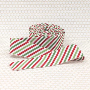 May include: A spool of fabric ribbon featuring a repeating diagonal stripe pattern in vibrant red, forest green, and white. The ribbon is wound into a tight cylinder with one end extending outward, showcasing the consistent striped design throughout.