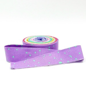 May include: A roll of colorful fabric strips with a purple strip in the foreground. The purple strip has a pattern of green stars and crescent moons. The roll of fabric is made up of various colors.