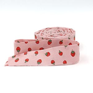 May include: A roll of pink fabric with a repeating pattern of red strawberries with green leaves. The fabric is unrolled in a loop, showing the pattern clearly. The fabric is likely used for sewing or crafting projects.
