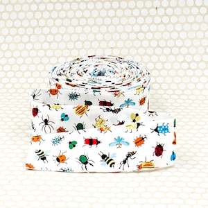 May include: A roll of white fabric ribbon featuring a repeating pattern of colorful insects including ladybugs, bees, ants, and beetles in shades of red, yellow, blue, green, and black. The ribbon is coiled into a flat, circular spool shape.