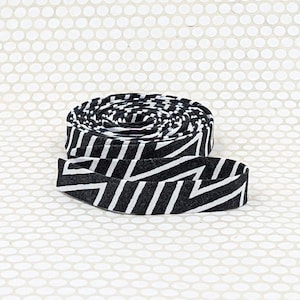 May include: A coiled strip of fabric tape featuring a repeating black and white geometric pattern of diagonal zig-zag lines. The textile ribbon is wound into a circular shape with one end extending outward, displaying the consistent graphic print.