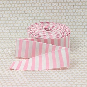 May include: A roll of fabric ribbon featuring a repeating pattern of vertical light pink and white stripes. The ribbon is wound into a tight circular coil with one end extending outward, displaying the consistent striped design across the surface.
