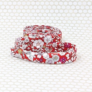 May include: A coiled strip of fabric featuring a vibrant red background with a repeating pattern of white, pink, and yellow flowers and green leaves. The textile ribbon is wound into a circular shape, displaying the floral print across its surface.