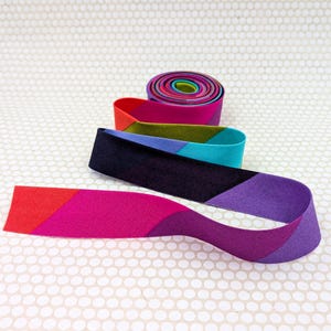 May include: A continuous roll of fabric tape featuring a vibrant, multi-colored diagonal pattern. The strip displays alternating sections of red, magenta, purple, black, teal, and olive green, creating a bold, geometric, and colorful design.