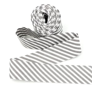 May include: A roll of fabric ribbon featuring a repeating pattern of diagonal gray and white stripes. The ribbon is wound into a tight circular spool at the top, with a long length of the striped material extending outward and curving across the frame.