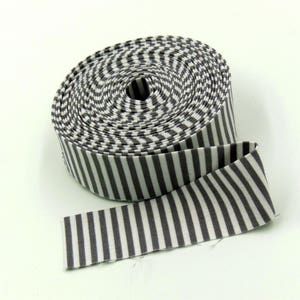 May include: A roll of fabric ribbon featuring a repeating pattern of vertical dark gray and white stripes. The ribbon is wound into a tight circular coil with one end extending outward, displaying the consistent striped design throughout the length.