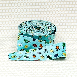 May include: A roll of light blue fabric ribbon featuring a repeating pattern of small, colorful insects including beetles, spiders, and bees in shades of red, yellow, green, and black, with a smooth texture and a finished edge on the ribbon strip.