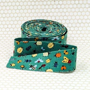 May include: A roll of teal fabric ribbon featuring a repeating pattern of various insects including beetles, ants, bees, and spiders in shades of yellow, orange, blue, and black. The ribbon is coiled into a circular shape with one end extending outward.