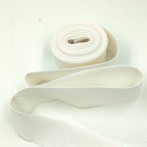 May include: A roll of off-white elastic band featuring a smooth, woven texture. The elastic is coiled into a circular shape at the top, with a long, flat strip extending downward, showcasing the consistent width and flexible material construction.