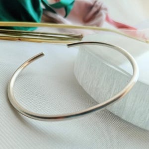 May include: A silver bangle bracelet with a smooth, rounded surface. The bracelet is open and has a simple, minimalist design.