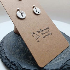 Può includere: Orecchini a lobo in argento con un design ovale martellato, ciascuno con un simbolo runico nero. Gli orecchini sono esposti su una scheda marrone con il testo "WildRaven925 hand crafted silver jewellery".