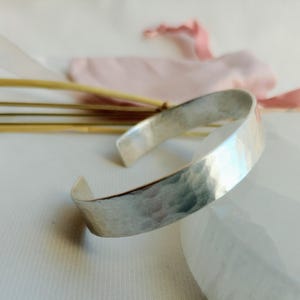 May include: A silver hammered bangle bracelet. The bracelet is made of a thick, flat band of metal that has been hammered to create a textured surface.