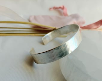 May include: A silver hammered bangle bracelet. The bracelet is made of a thick, flat band of metal that has been hammered to create a textured surface.