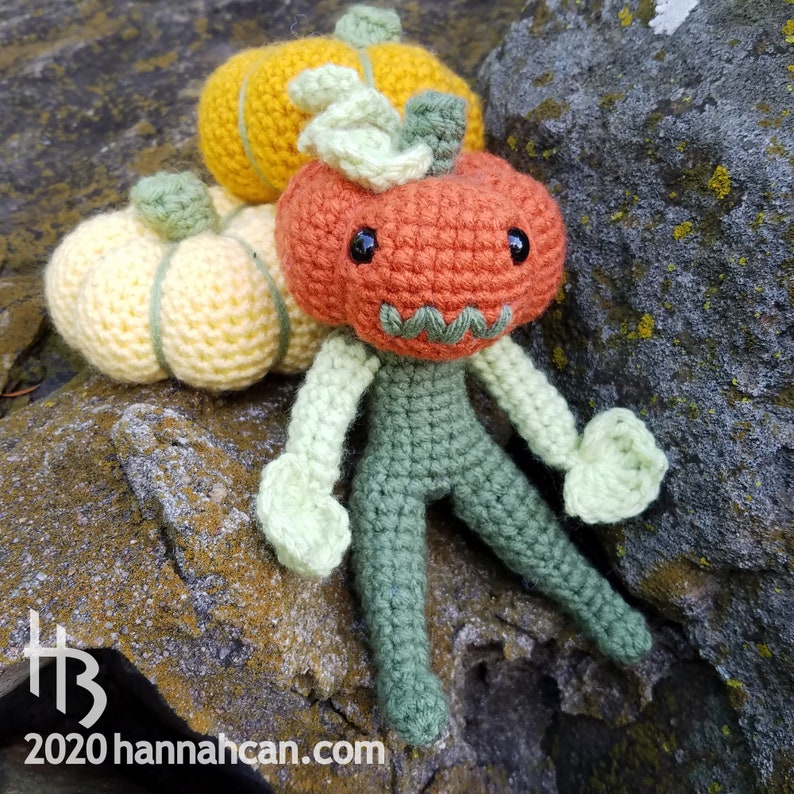 May include: A crocheted pumpkin character with a green body and orange head. The character is sitting on a rock with two smaller crocheted pumpkins beside it.