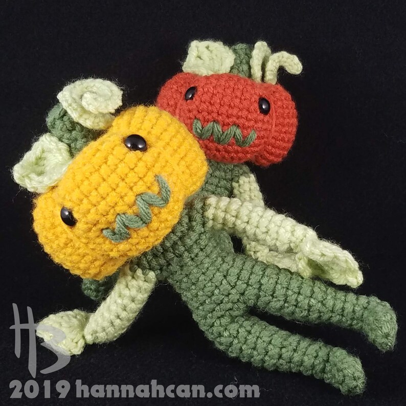 May include: Two crocheted pumpkin plushies, one green and one yellow, with black eyes and stitched mouths. The green pumpkin has a red pumpkin head with a stitched face.