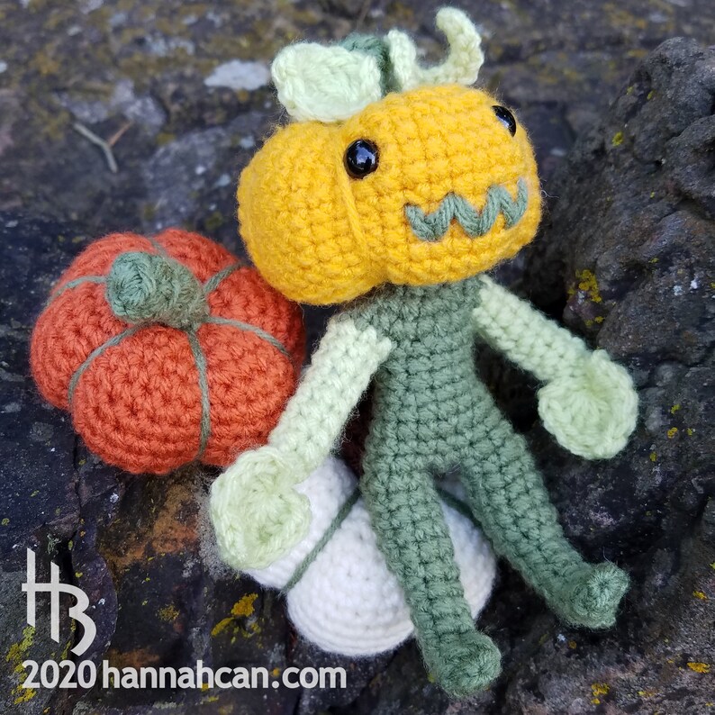 May include: A crocheted pumpkin character with a green body and a yellow pumpkin head. The character is sitting on a white crocheted ball and holding a small orange crocheted pumpkin.
