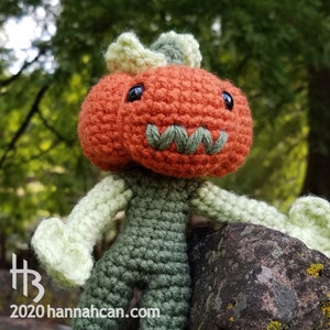 May include: A crocheted pumpkin character with a green body and a bright orange pumpkin head. The character has a stitched-on smile and black eyes. The character is sitting on a rock with green moss.
