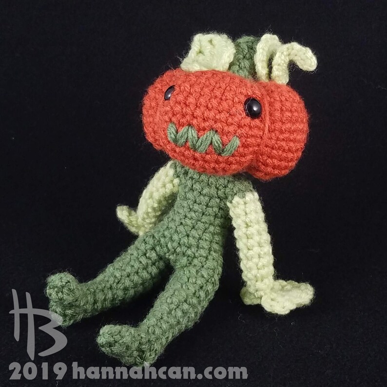 May include: A crocheted pumpkin-headed creature with green arms and legs. The creature has a stitched-on face with a green mouth and black eyes.