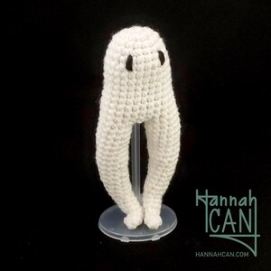 May include: White crocheted ghost with black eyes standing on a clear plastic stand. The ghost has long, thin legs. The stand has a round base. The text "Hannah CAN" is visible in the lower right corner of the image.
