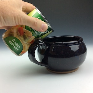 May include: A hand pouring chicken with rice soup from a can into a dark blue ceramic bowl with a handle.