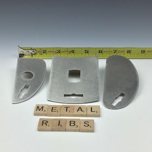 May include: Three metal pieces with different shapes and cutouts. The pieces are arranged on a white surface with a yellow measuring tape and letter tiles spelling "METAL" and "RIBS".