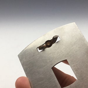May include: A silver metal plate with a cut-out shape and a small, rusty metal screw in the center.